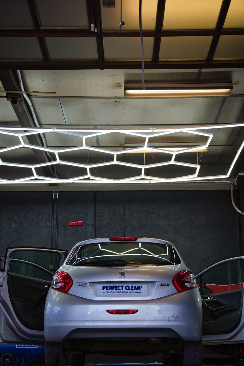 Vue intérieure de l'atelier : Peugeot 208 centrée sous la rampe hexagonale LED, portières arrière ouvertes en symétrie