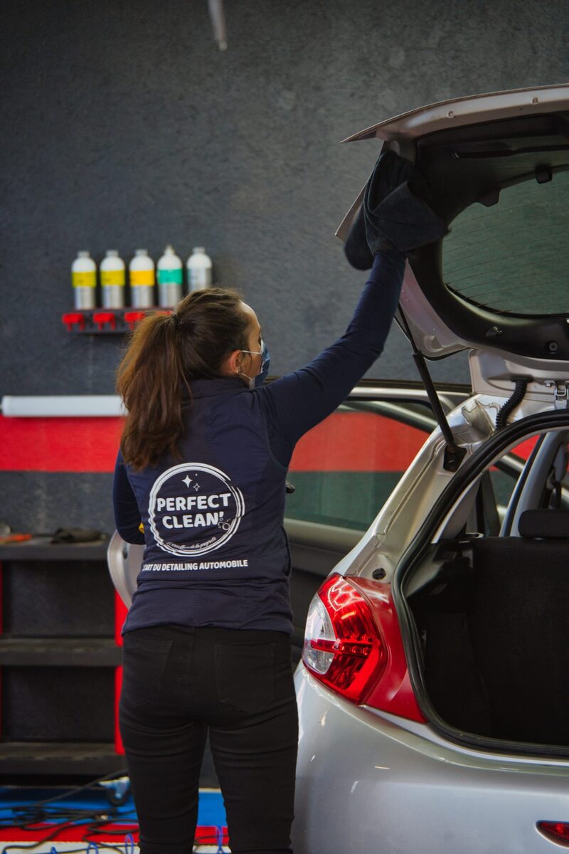Détailleuse de dos essuyant le hayon, logo 'L'art du detailing automobile' visible sur la veste